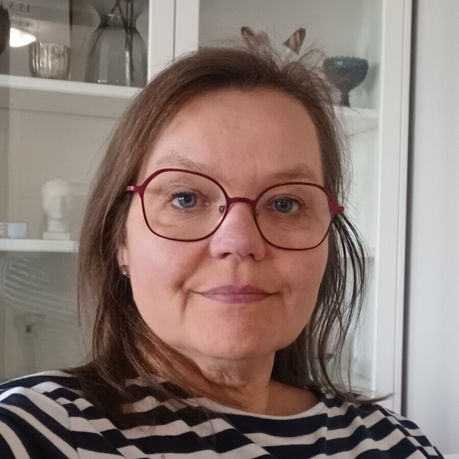 Nainen, jolla on olkapäiden pituiset ruskeat hiukset ja punasankaiset silmälasit, ja jolla on yllään mustavalkoinen raidallinen paita, istuu sisätiloissa valkoisen kaapin edessä, jossa on hyllyjä ja koriste-esineitä.