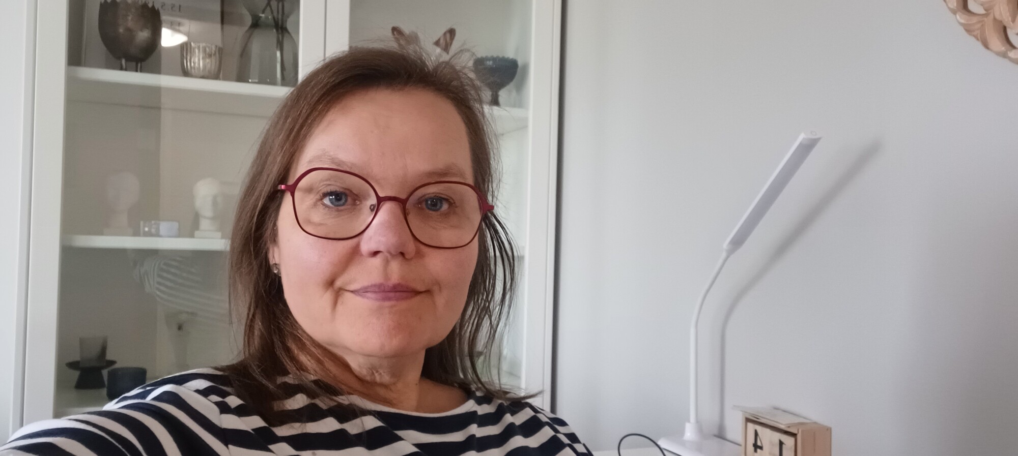 Nainen, jolla on olkapäiden pituiset ruskeat hiukset ja punasankaiset silmälasit, ja jolla on yllään mustavalkoinen raidallinen paita, istuu sisätiloissa valkoisen kaapin edessä, jossa on hyllyjä ja koriste-esineitä.