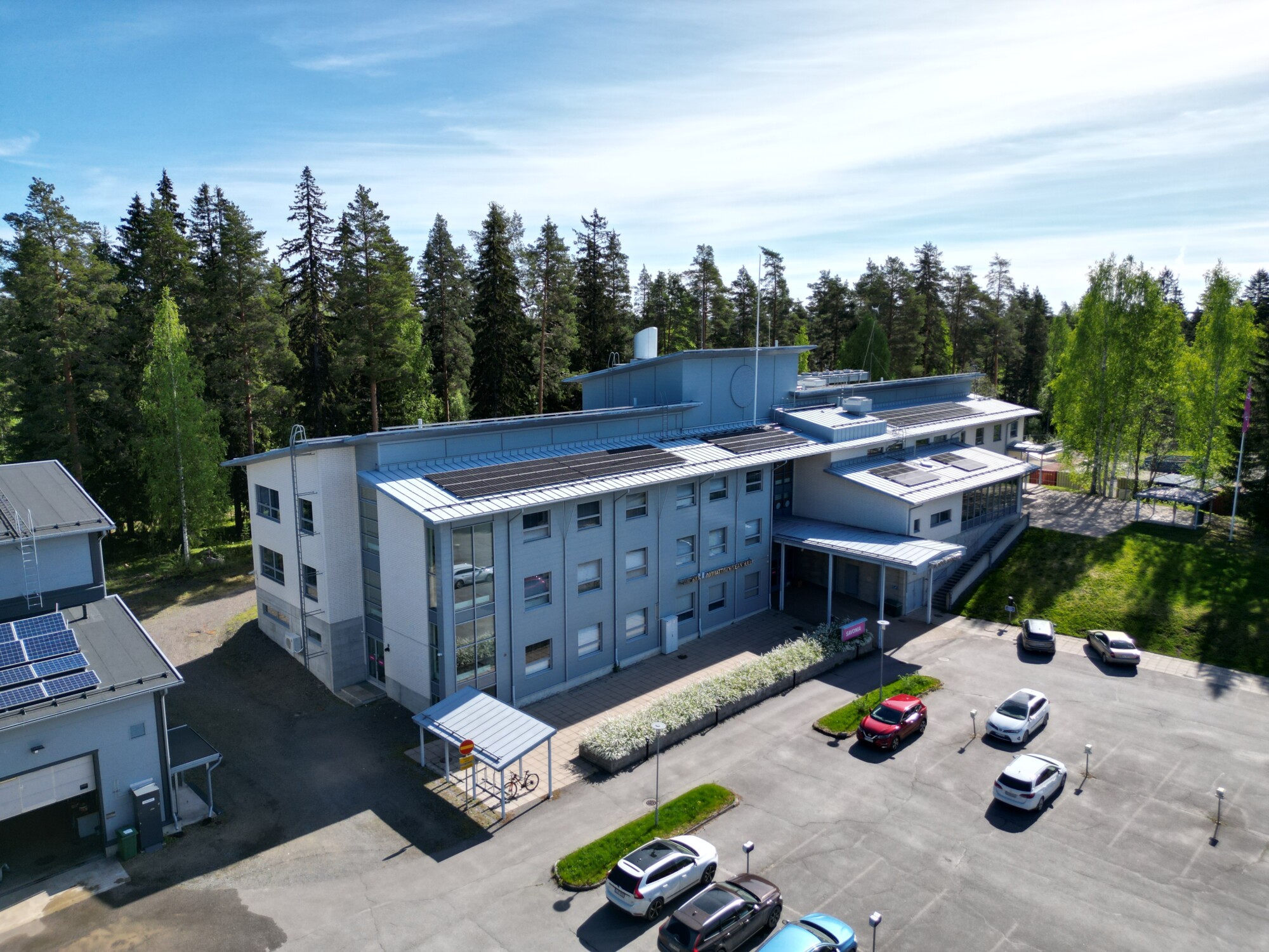 Moderni, vaalea rakennus, jonka katolla on aurinkopaneelit, on puiden ympäröimä. Viereiselle parkkipaikalle on pysäköity useita autoja, ja taivas on kirkas ja aurinkoinen.