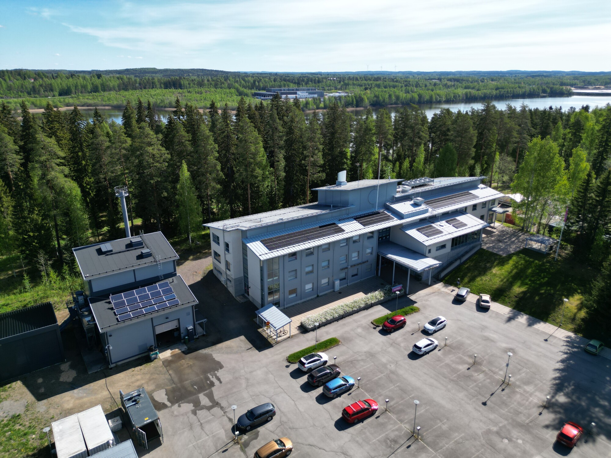 Ilmakuva modernista rakennuskompleksista, jonka katoilla on aurinkopaneeleita, puiden ympäröimänä ja parkkipaikalla on useita autoja. Taustalla on joki ja metsäinen maisema sinisen taivaan alla.