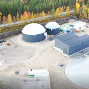 Ilmakuva biokaasulaitoksesta, jossa on pyöreitä varastosäiliöitä ja suorakaiteen muotoisia rakennuksia, syksynväristen puiden ja läheisen tien ympäröimänä.