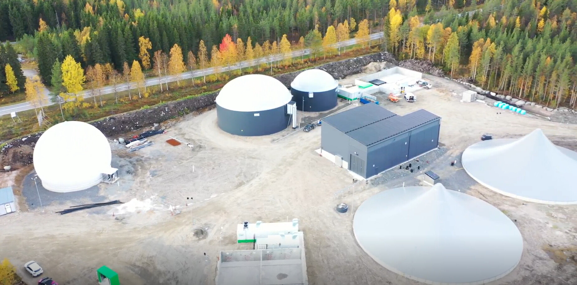 Ilmakuva biokaasulaitoksesta, jossa on pyöreitä varastosäiliöitä ja suorakaiteen muotoisia rakennuksia, syksynväristen puiden ja läheisen tien ympäröimänä.