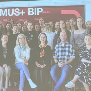 Noin 25 hengen ryhmä, enimmäkseen naisia, poseeraa yhdessä sisätiloissa punavalkoisen ERASMUS+ BIP 2026 -banderollin edessä. Suurin osa seisoo, kuusi ihmistä istuu edessä.