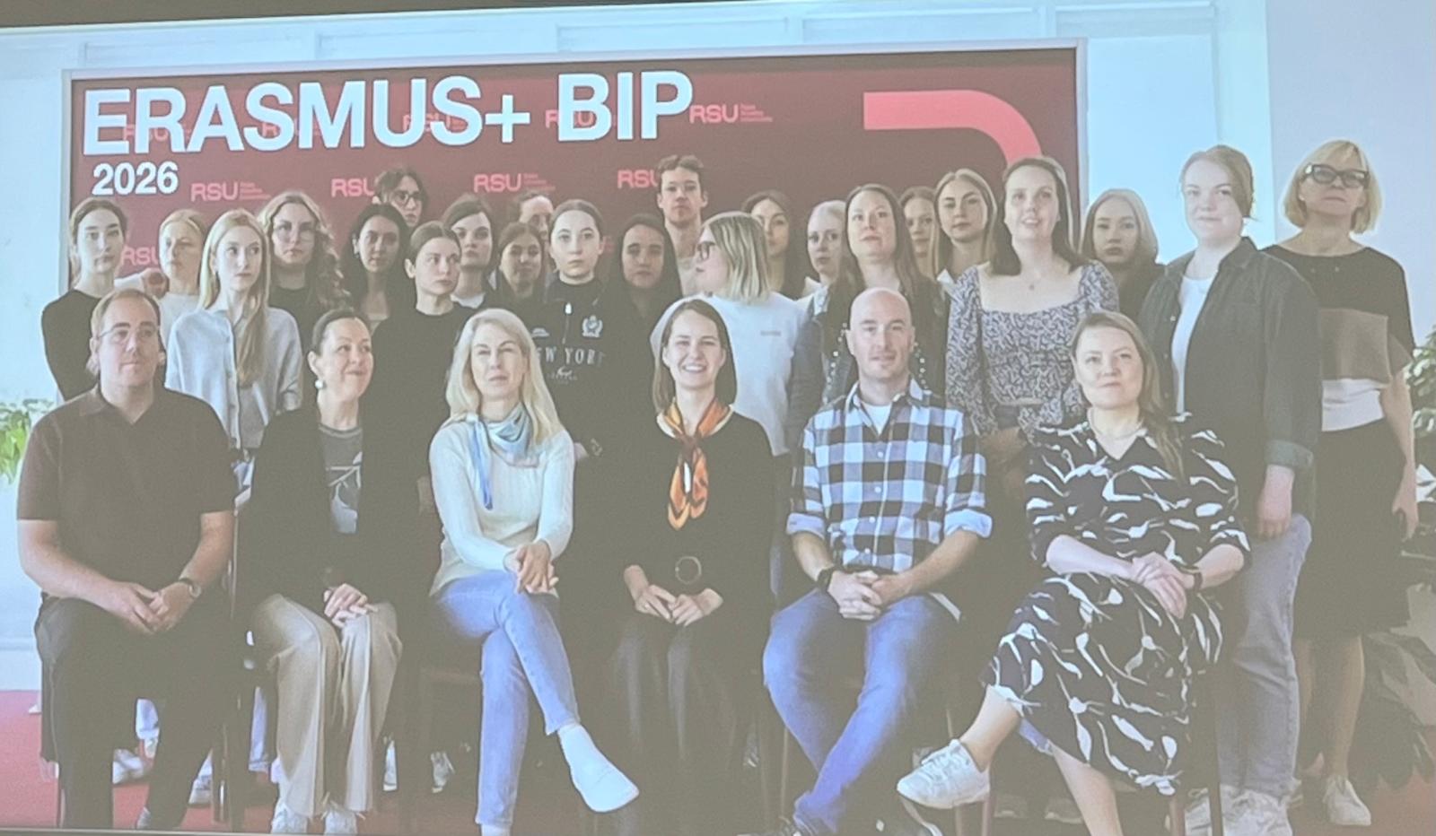 Noin 25 hengen ryhmä, enimmäkseen naisia, poseeraa yhdessä sisätiloissa punavalkoisen ERASMUS+ BIP 2026 -banderollin edessä. Suurin osa seisoo, kuusi ihmistä istuu edessä.