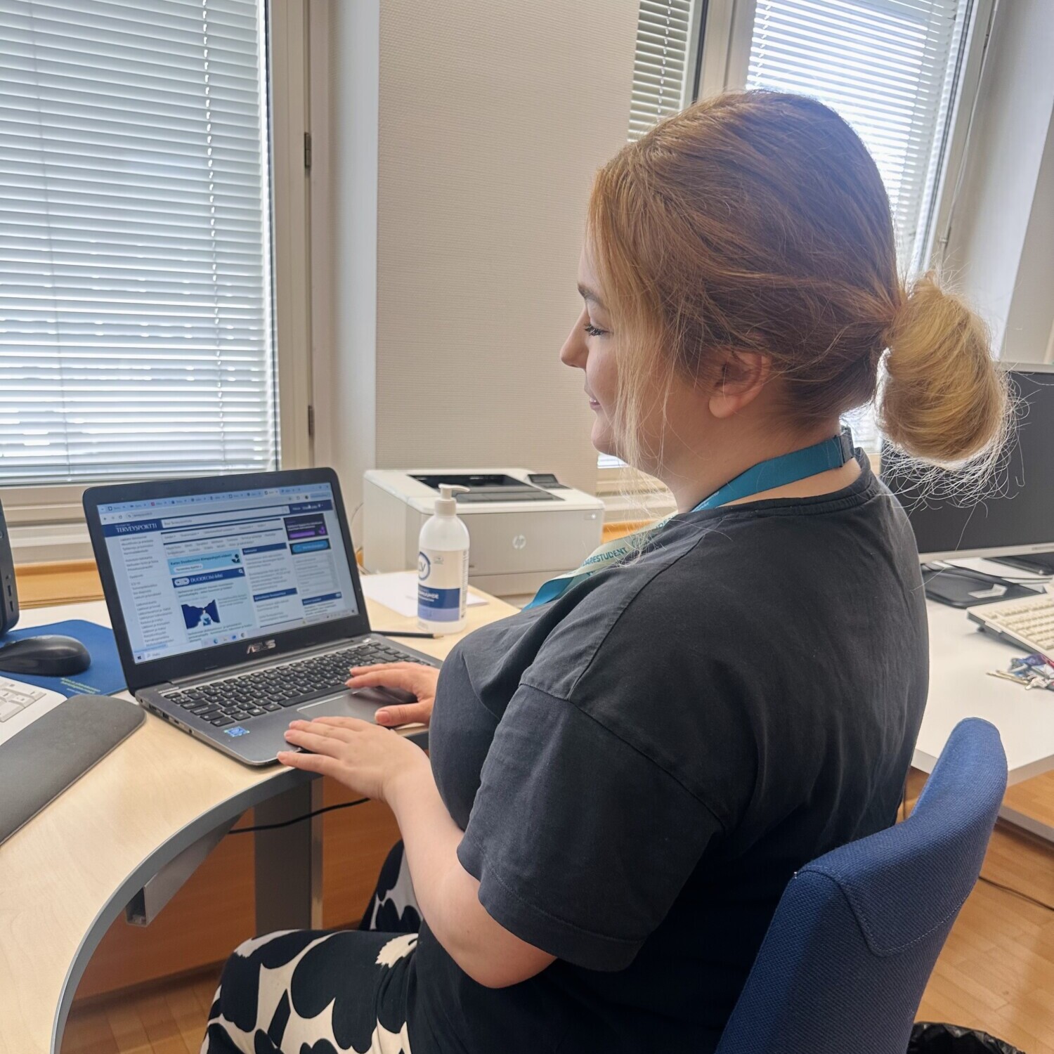 Nainen, jolla on punaiset hiukset poninhännässä, istuu työpöydän ääressä ja kirjoittaa kannettavalla tietokoneella. Hänellä on yllään musta t-paita ja kuviolliset housut. Työpöydällä on toimistotarvikkeita, tulostin ja desinfiointiainepullo; taustalla on ikkunoita.
