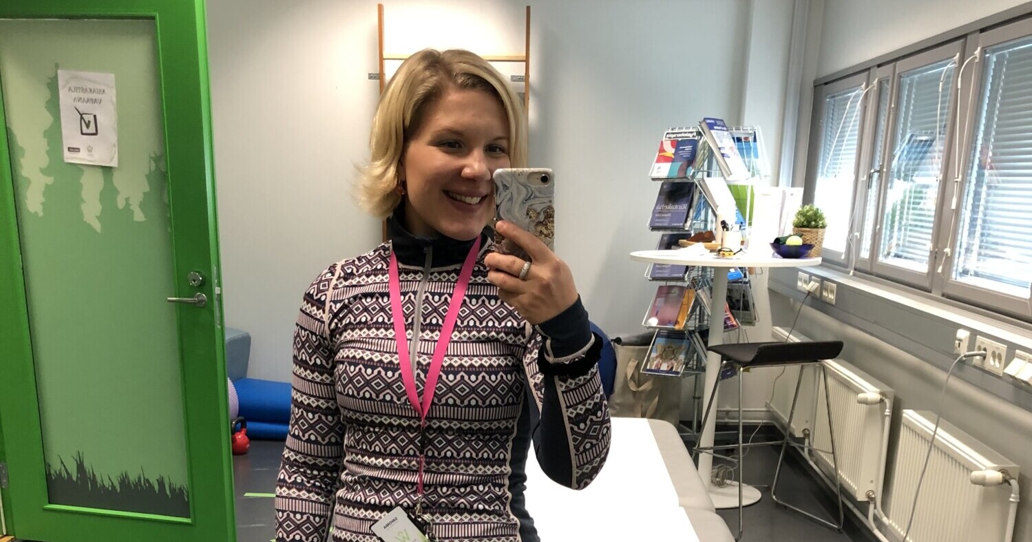 Lyhyet vaaleat hiukset omaava nainen, jolla on kuvioitu pusero ja vaaleanpunainen kaulanauha, ottaa selfien valoisassa, modernissa toimistossa, jossa on suuret ikkunat, lehtiä hyllyillä ja vihreä ovi taustalla.