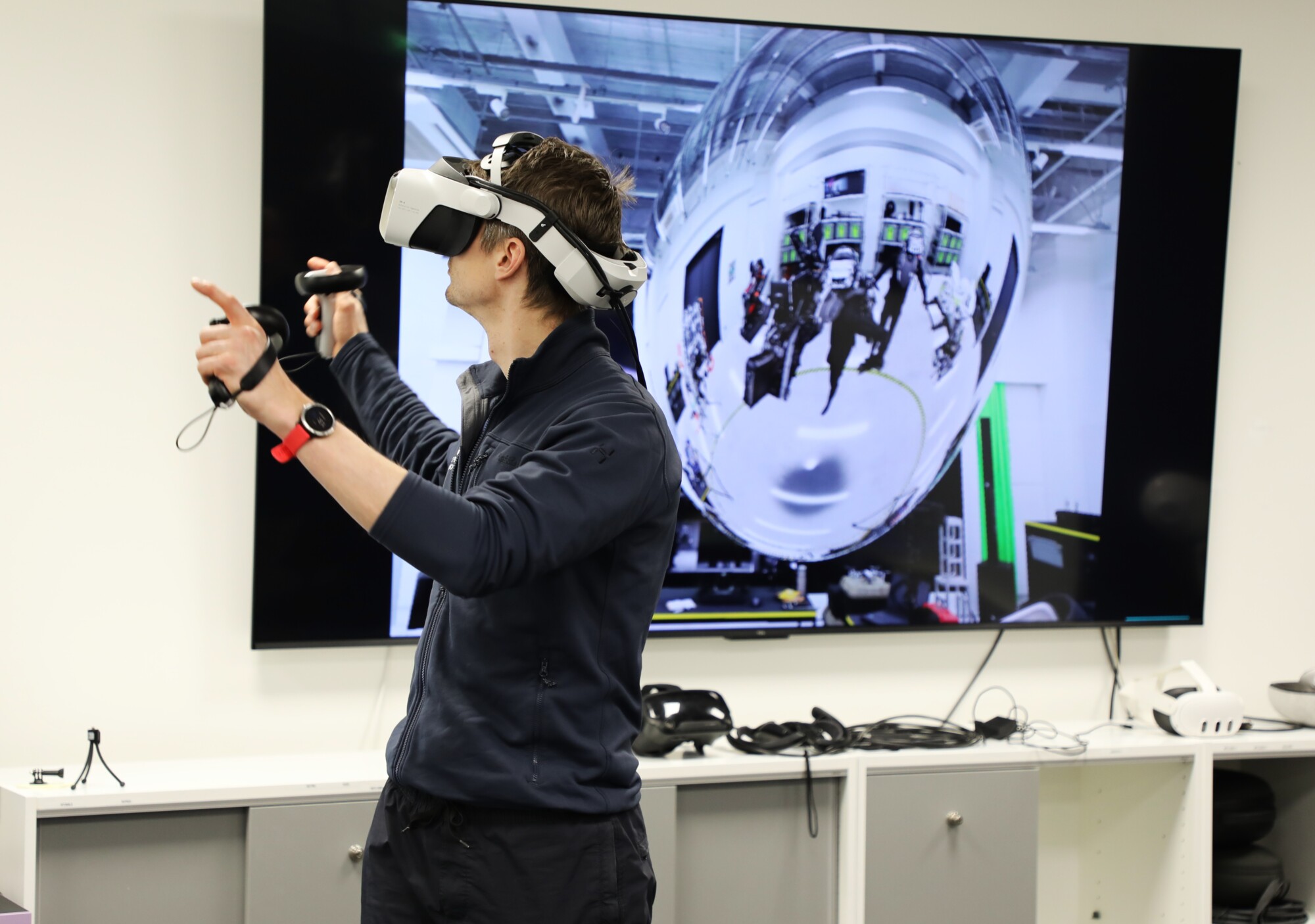 Henkilö, jolla on virtuaalitodellisuuskuulokkeet ja ohjaimet kädessään, on vuorovaikutuksessa virtuaalitodellisuusympäristön kanssa, kun hänen takanaan olevalla suurella näytöllä näkyy vääristynyt kuva, joka mahdollisesti näyttää, mitä hän näkee virtuaalitodellisuudessa.