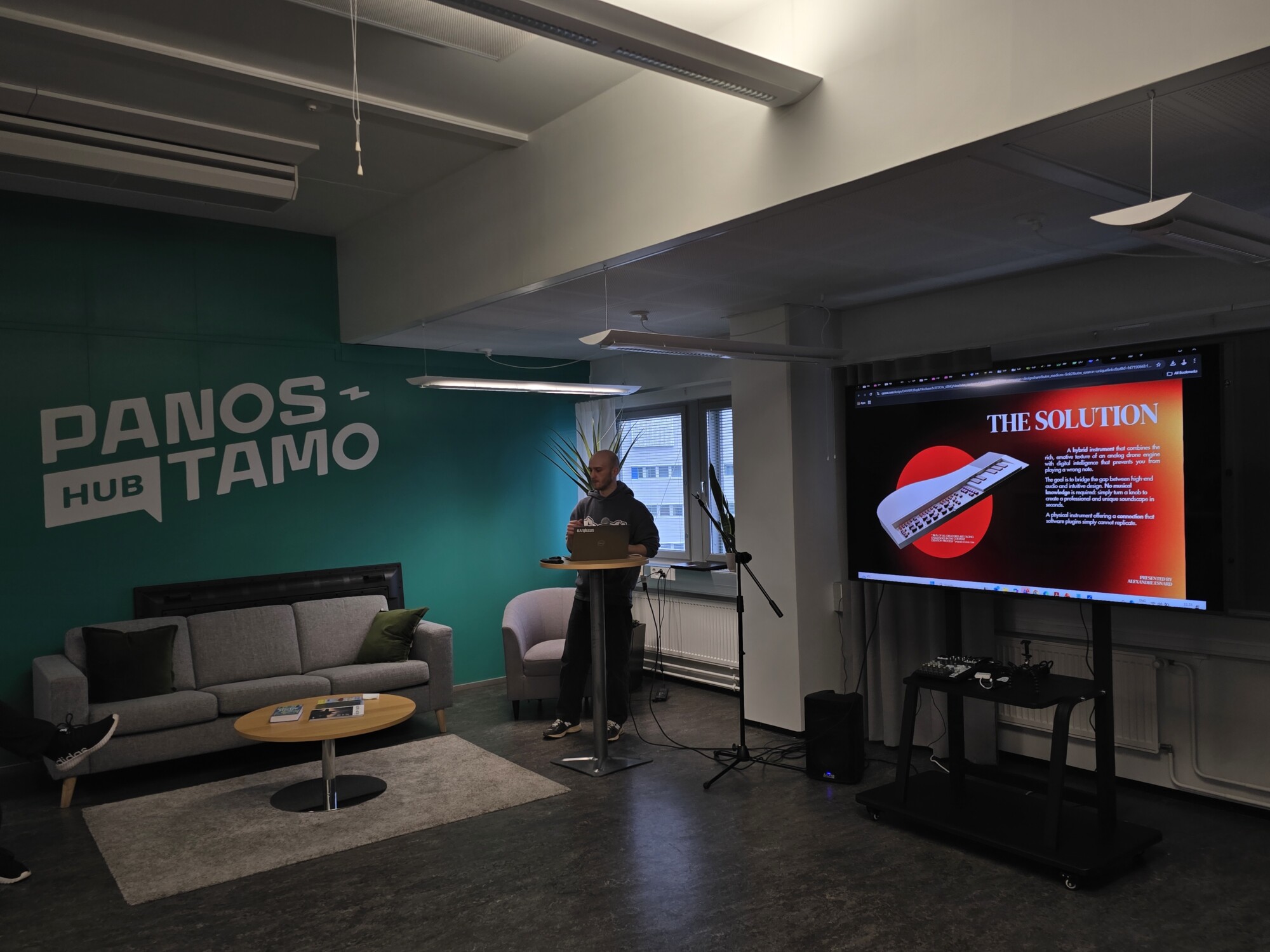 Henkilö seisoo korokkeella modernissa toimistotilassa pitämässä esitelmää. Suurella näytöllä näkyy dia otsikolla THE SOLUTION, jossa on tekstiä ja grafiikkaa. Takana olevalla vihreällä seinällä lukee PANOS HUB TAMO.