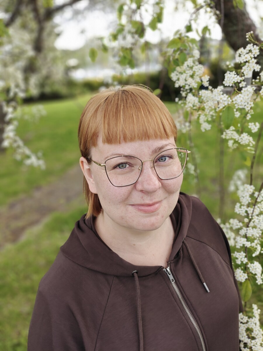 Henkilö, jolla on lyhyet punaiset hiukset, silmälasit ja ruskea huppari, seisoo ulkona valkoisten kukkivien oksien vieressä ja hymyilee kameralle. Taustalla on vihreää ruohoa ja puita.