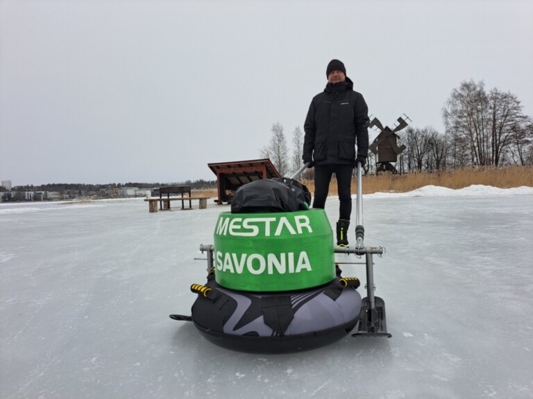 Mustiin talvivaatteisiin pukeutunut henkilö seisoo jäätyneellä järvellä vihreän ja mustan jääveneen vieressä, jossa lukee MESTAR SAVONIA. Taustalla näkyy paljaita puita ja puinen tuulimylly.