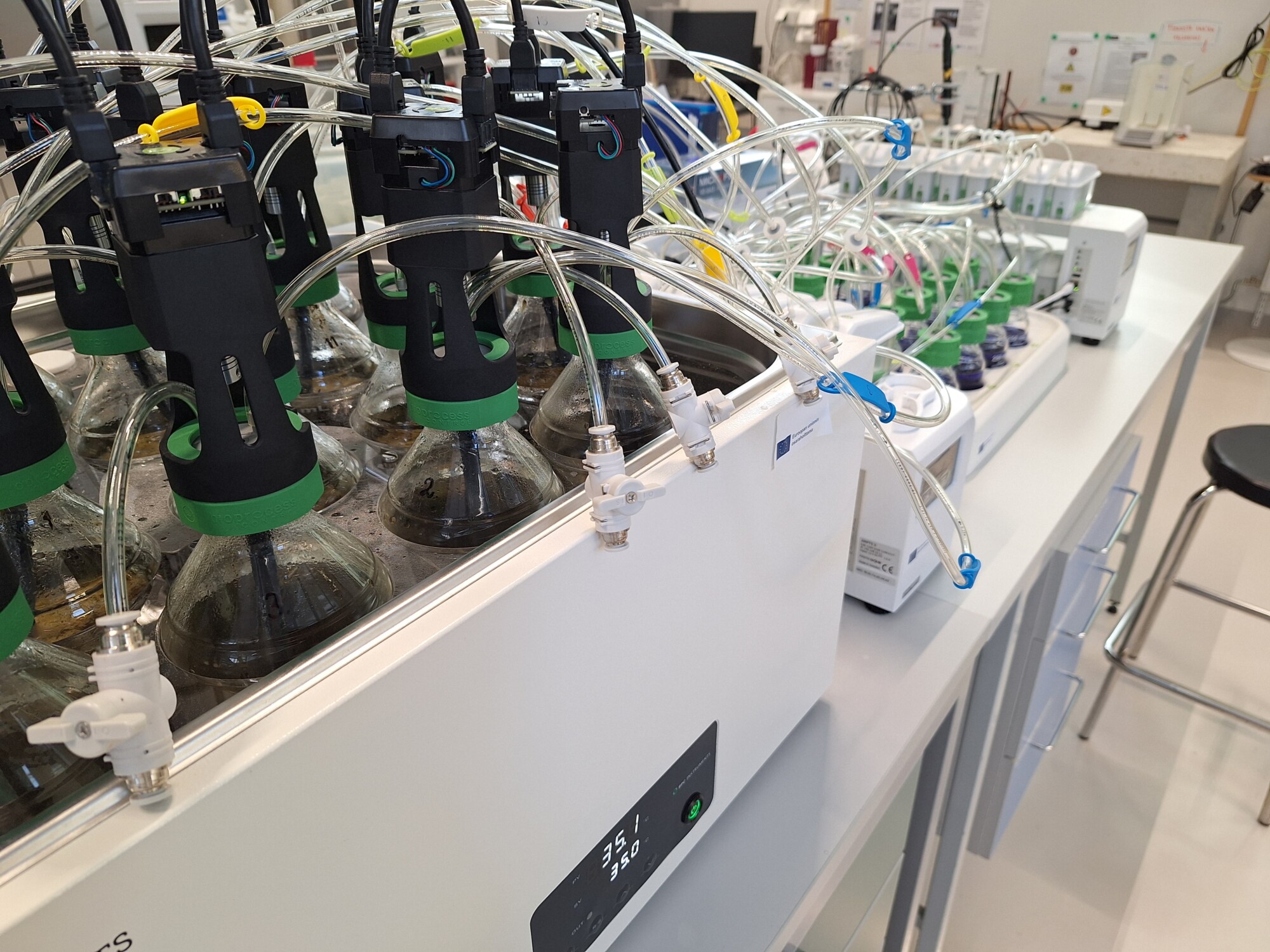Laboratorioasetelma, jossa on lasipulloja, jotka on liitetty putkiin ja elektronisiin laitteisiin, todennäköisesti kemiallisia tai biologisia kokeita varten, ja joka on sijoitettu tiskipöydälle, jossa on digitaalisia säätimiä ja erilaisia pulloja taustalla.