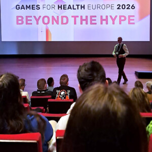 Puhuja seisoo lavalla konferenssin yleisön edessä. Hänen takanaan olevalla näytöllä näkyy suurilla kirjaimilla GAMES FOR HEALTH EUROPE 2026 BEYOND THE HYPE.