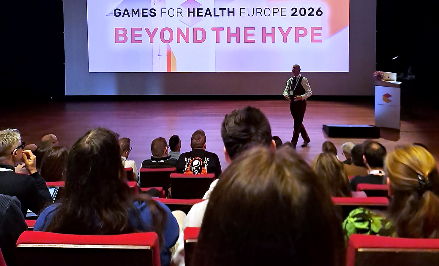Puhuja seisoo lavalla konferenssin yleisön edessä. Hänen takanaan olevalla näytöllä näkyy suurilla kirjaimilla GAMES FOR HEALTH EUROPE 2026 BEYOND THE HYPE.