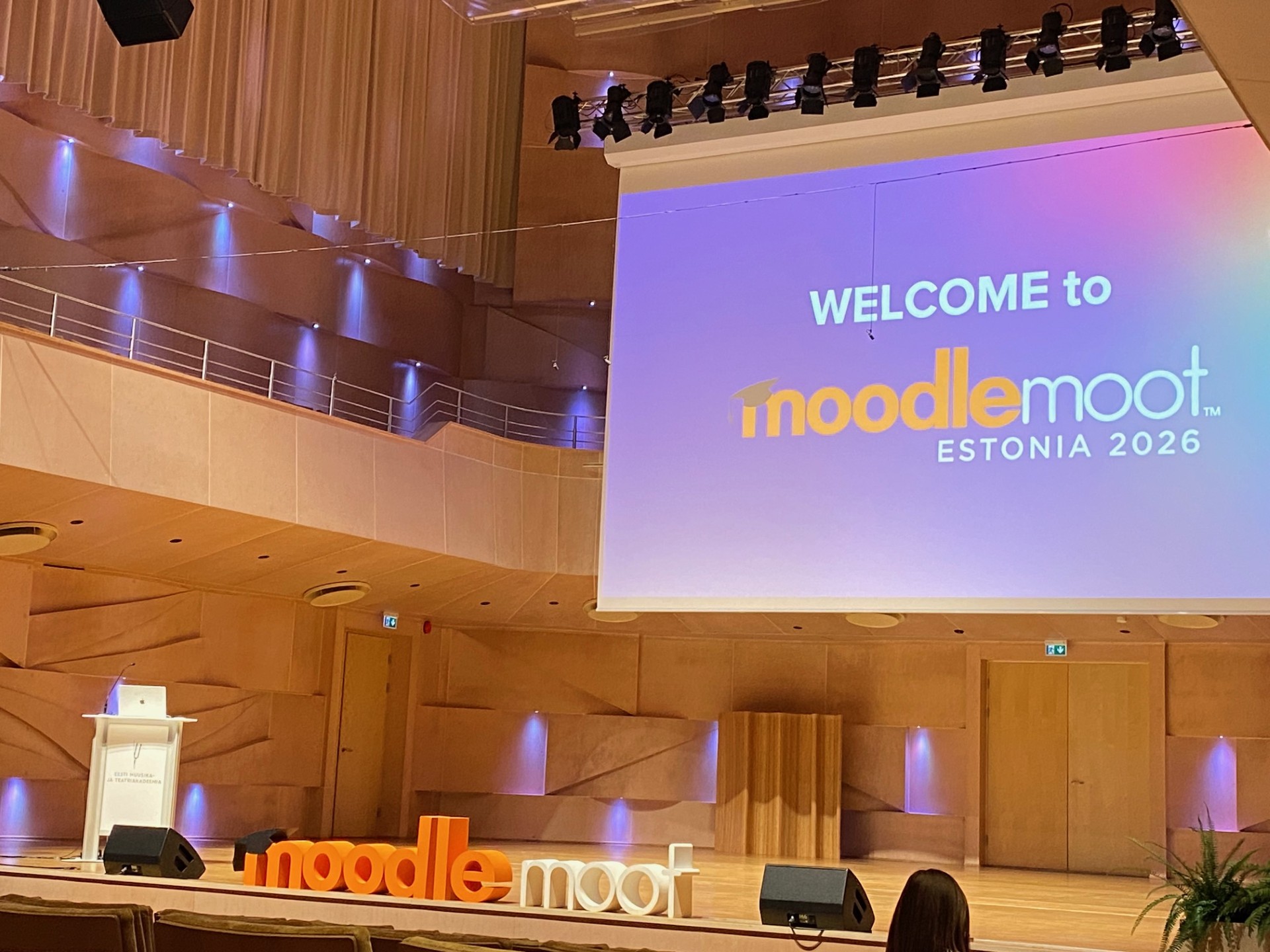 Moderni auditorio, jonka näytöllä lukee "Tervetuloa MoodleMoot Estonia 2026". Lavalla on koroke, suuret koristeelliset kirjaimet, joissa lukee "noodlemoot", ja lämmin puupanelointi.