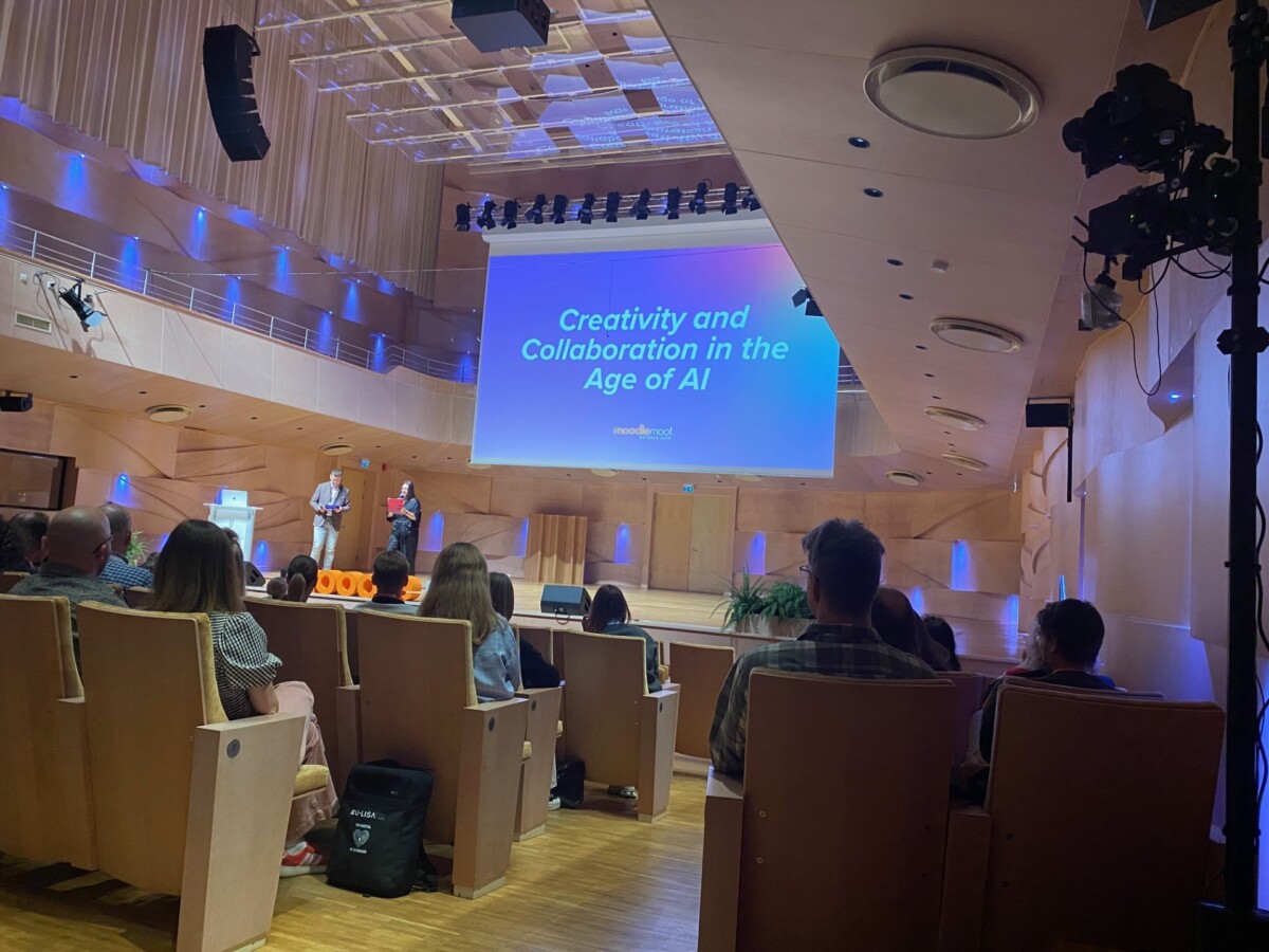 Konferenssiyleisö istuu lavaa vastapäätä, jossa kaksi esittelijää seisoo modernissa auditoriossa näytön vieressä, jossa näkyy teksti Creativity and Collaboration in the Age of AI.