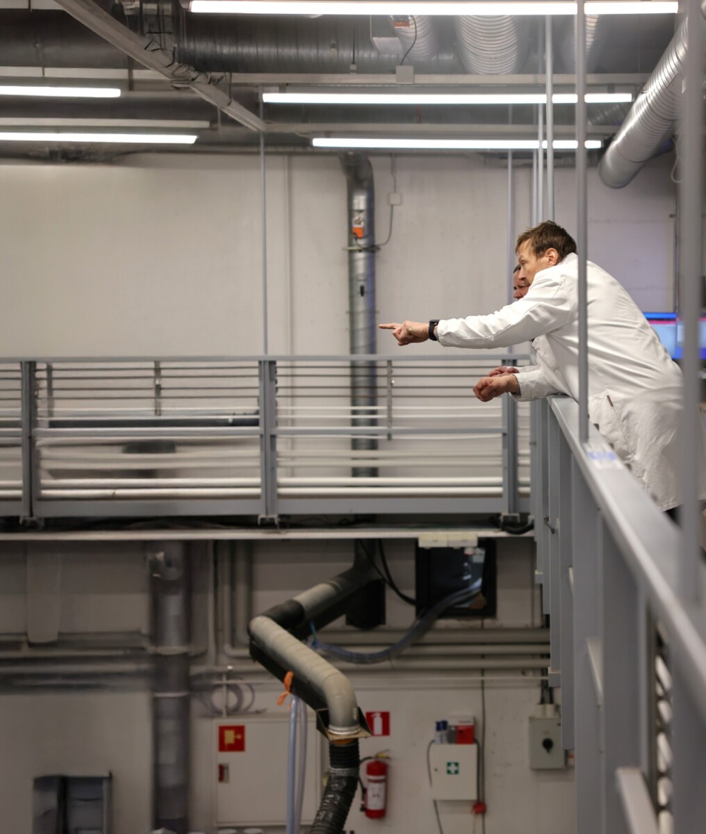 Kaksi valkoisiin laboratoriotakkeihin pukeutunutta ihmistä nojaa teollisuuslaitoksen ylemmällä tasolla olevan kaiteen yli, ja toinen osoittaa kohti jotakin alla olevaa. Taustalla näkyy suuria putkia ja teollisuuslaitteita.