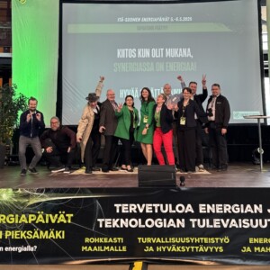 Joukko ihmisiä poseeraa ja hymyilee lavalla suomenkielistä tekstiä sisältävän näytön edessä energia-aiheisessa tapahtumassa. Lavan banneri mainostaa Pieksämäellä toukokuussa 2026 järjestettävää energiakonferenssia.