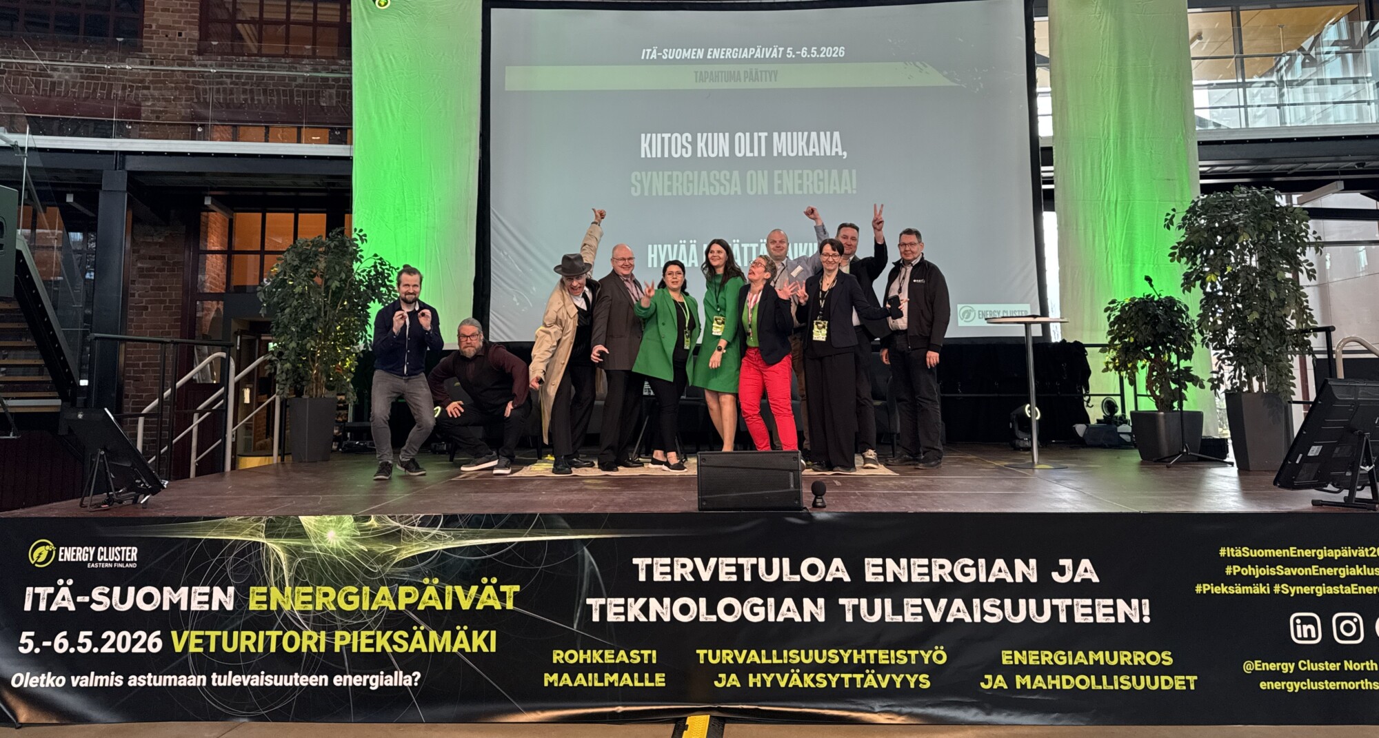 Joukko ihmisiä poseeraa ja hymyilee lavalla suomenkielistä tekstiä sisältävän näytön edessä energia-aiheisessa tapahtumassa. Lavan banneri mainostaa Pieksämäellä toukokuussa 2026 järjestettävää energiakonferenssia.