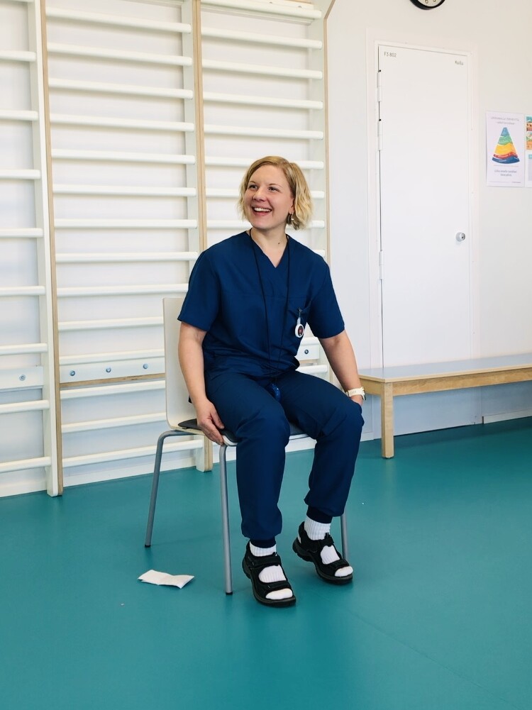 Sinisiin hoitohousuihin pukeutunut henkilö istuu tuolilla valoisassa huoneessa, jossa on kuntosalilaitteita, ja hymyilee. Heillä on sidotut saappaat ja raidalliset sukat. Heidän takanaan olevalla valkoisella seinällä on kello ja värikäs juliste.