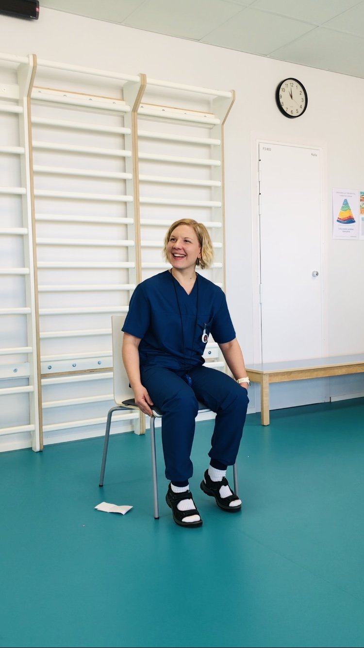 Sinisiin hoitohousuihin pukeutunut henkilö istuu tuolilla valoisassa huoneessa, jossa on kuntosalilaitteita, ja hymyilee. Heillä on sidotut saappaat ja raidalliset sukat. Heidän takanaan olevalla valkoisella seinällä on kello ja värikäs juliste.