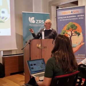Mies esiintyy puhujakorokkeella, jonka näytöllä on lannoitteita kuvaavia kaavioita ja kuvia. Hänen edessään istuu kaksi naista, joista toinen katsoo kannettavaa tietokonetta.
