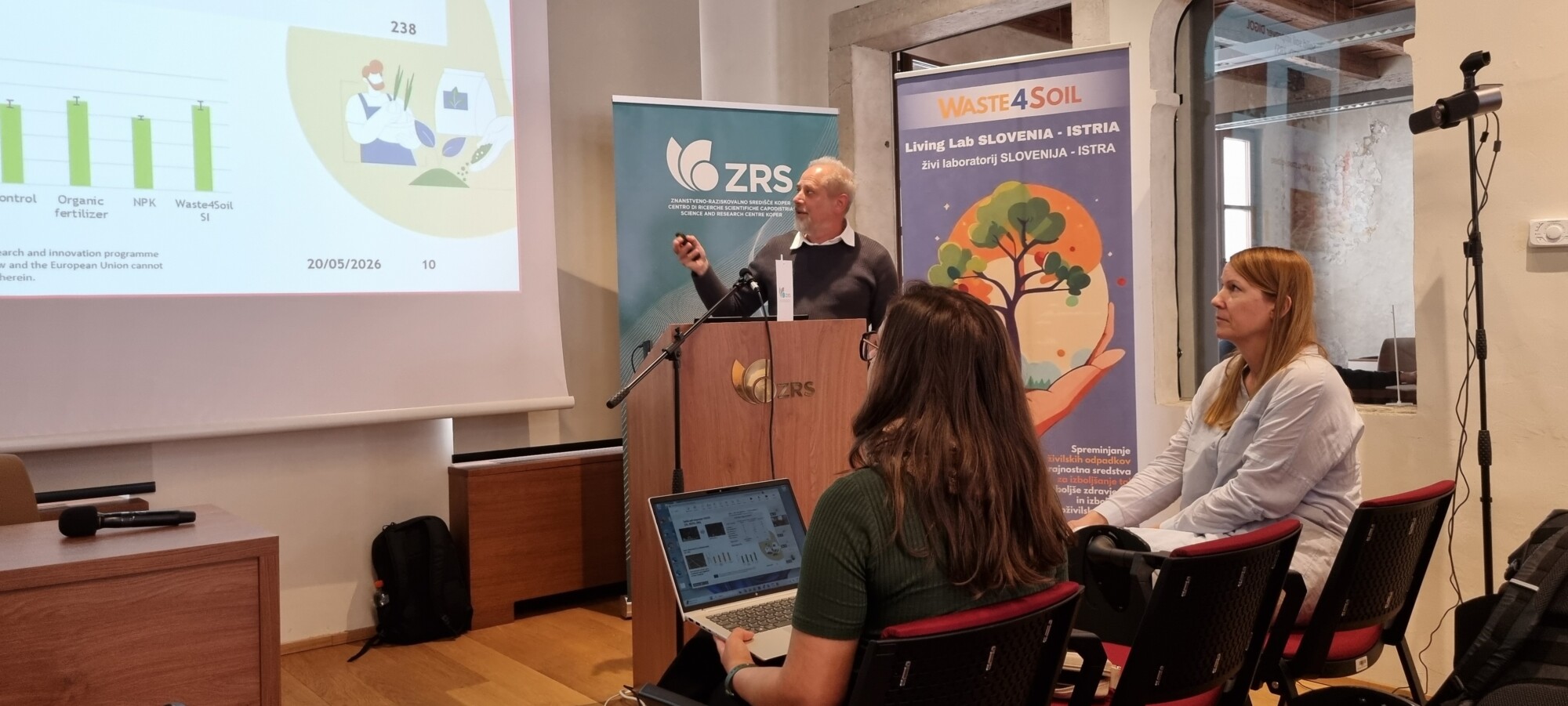 Mies esiintyy puhujakorokkeella, jonka näytöllä on lannoitteita kuvaavia kaavioita ja kuvia. Hänen edessään istuu kaksi naista, joista toinen katsoo kannettavaa tietokonetta.