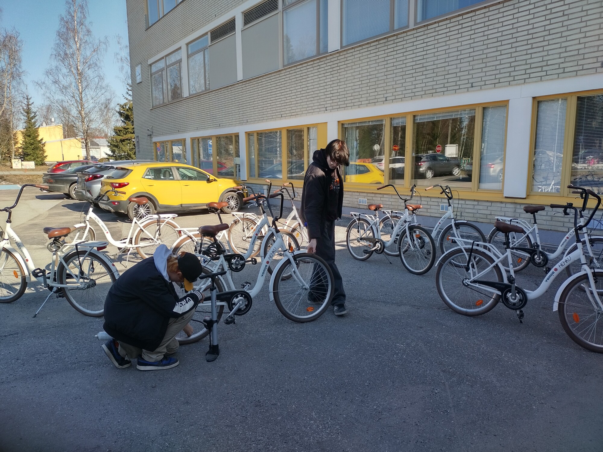 Kaksi ihmistä valmistelee valkoisia polkupyöriä rakennuksen ulkopuolella, jossa on suuret ikkunat. Toinen henkilö kyyristyy tarkastamaan pyörää, kun toinen seisoo lähellä. Taustalla on useita pysäköityjä pyöriä ja autoja.