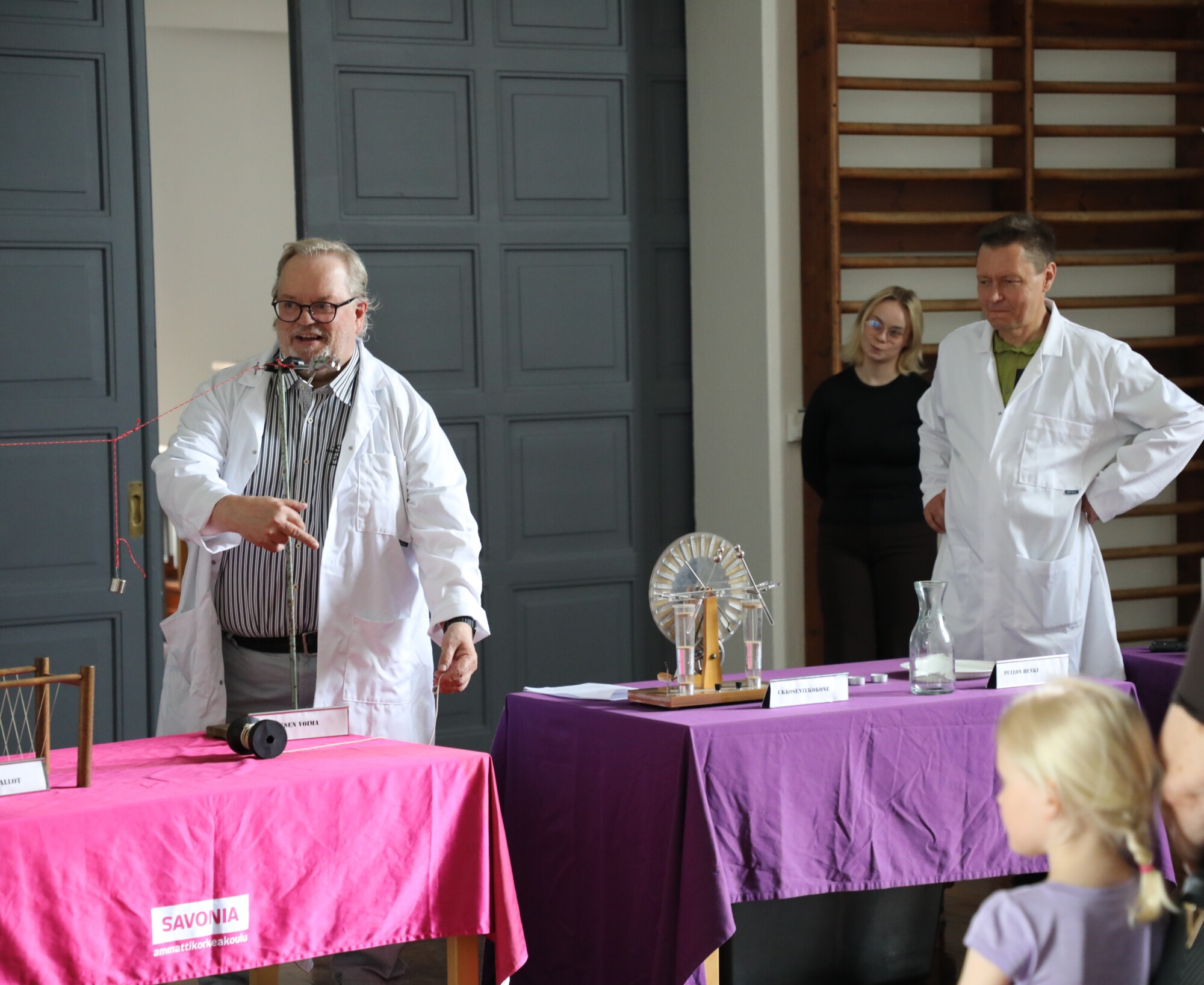Valkoiseen laboratoriotakkiin pukeutunut mies demonstroi tieteellistä koetta pöydällä, jossa on välineitä, kun toinen laboratoriotakkiin pukeutunut mies ja kaksi katsojaa katsovat. Etualalla istuu lapsi. Kohtaus on sisätiloissa.