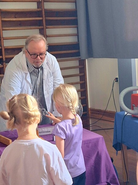 Vanhempi valkotakkinen mies demonstroi tieteellistä koetta kahdelle vaaleahiuksiselle nuorelle tytölle luokkahuoneessa. Tytöt tarkkailevat koetta violetilla päällystetyllä pöydällä ja ovat vuorovaikutuksessa sen kanssa.