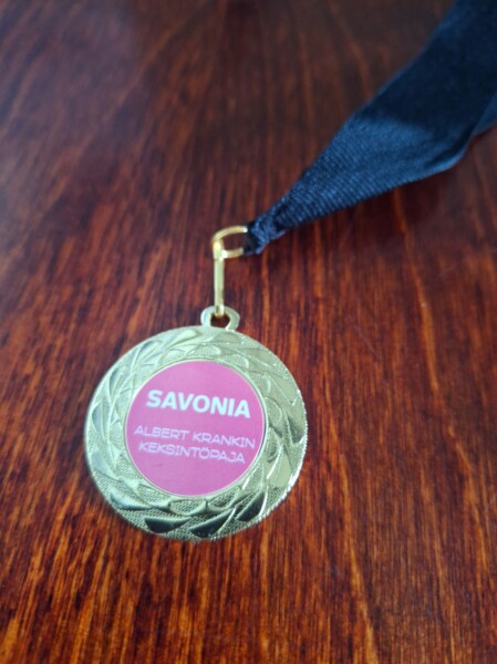 Kultainen mitali, jossa on musta nauha, makaa puupinnalla. Mitalin keskellä vaaleanpunaisella pohjalla lukee SAVONIA ALBERT KRANKIN KEKSINTÖPAJA.