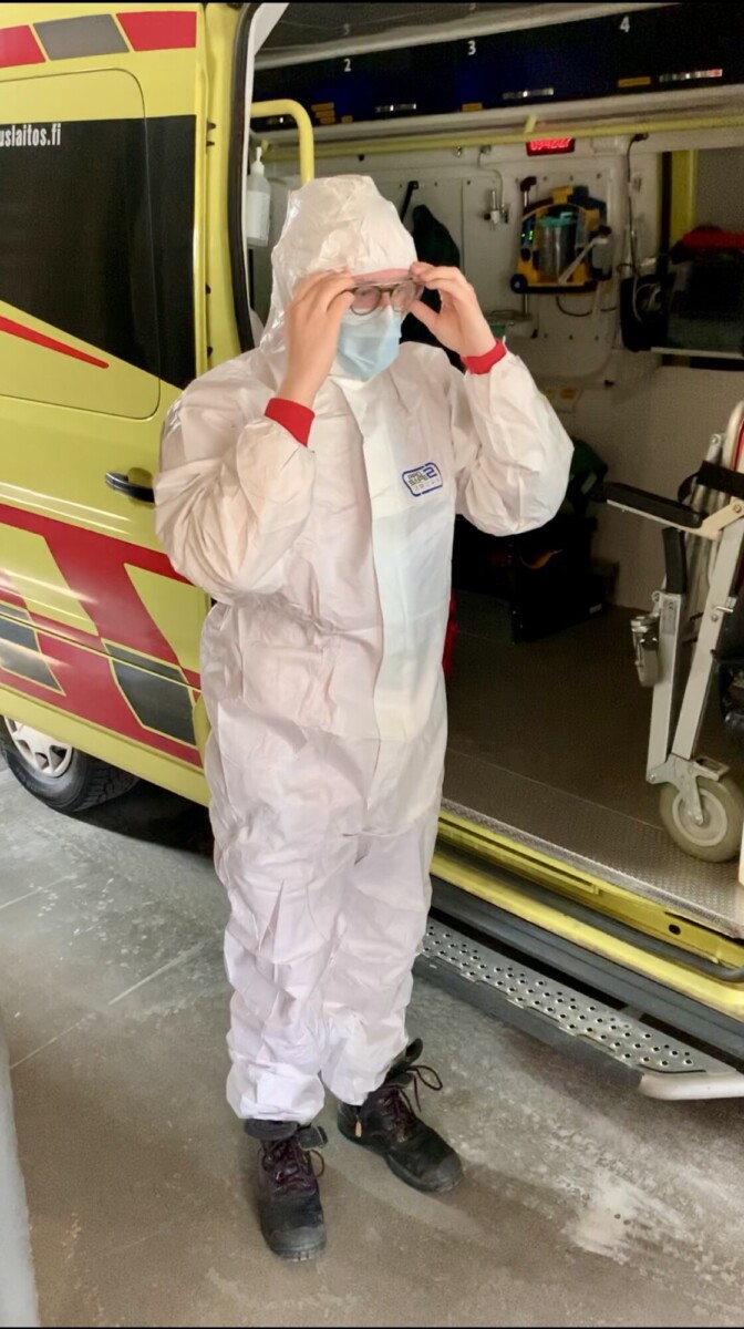 Henkilö, jolla on valkoinen suojapuku, kasvonaamari ja suojalasit, seisoo avoimen ambulanssin vieressä ja säätää silmälasejaan. Ambulanssin sisällä näkyy lääkinnällisiä laitteita.