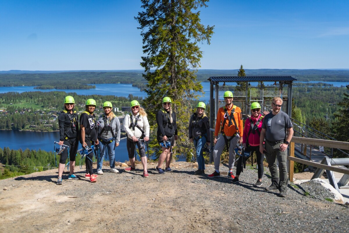 Ryhmä ihmisiä, joilla on kypärät ja valjaat, seisoo maisemallisella näköalapaikalla, josta on näkymät metsään, järviin ja siniselle taivaalle, valmistautumassa köysirataelämykseen.