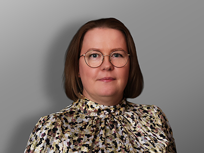 Nainen, jolla on suorat ruskeat hiukset, pyöreät silmälasit ja kukkakuvioinen pusero, on kameraan päin yksinkertaista harmaata taustaa vasten.