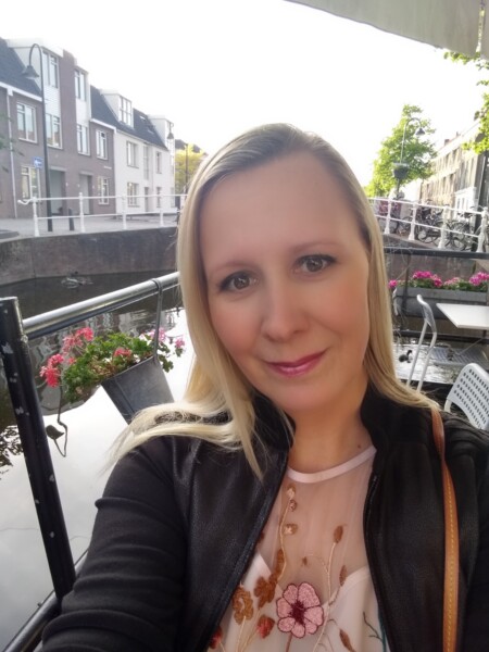 Nainen, jolla on pitkät vaaleat hiukset, ottaa selfien ulkona kanavan rannalla, taustalla kukkia, kaiteet ja rakennuksia. Hän hymyilee ja hänellä on yllään vaalea kukkatoppi ja musta takki.