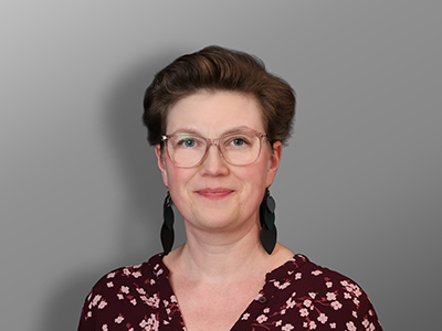 Jenny Robson, Nainen, jolla on lyhyet ruskeat hiukset, silmälasit ja tummat korvakorut, ja jolla on yllään ruskeankeltainen pusero, jossa on valkoinen kukkakuvio, hymyilee harmaan taustan edessä.