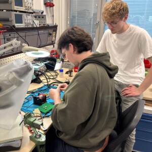 Elektroniikkatyöpisteellä työskentelee kaksi henkilöä. Toinen heistä, joka istuu ja jolla on yllään vihreä huppari, kokoaa piirikomponentteja, kun taas toinen, joka seisoo valkoisessa t-paidassa, seuraa työtä tarkasti. Heidän ympärillään näkyy työkaluja ja laitteita.