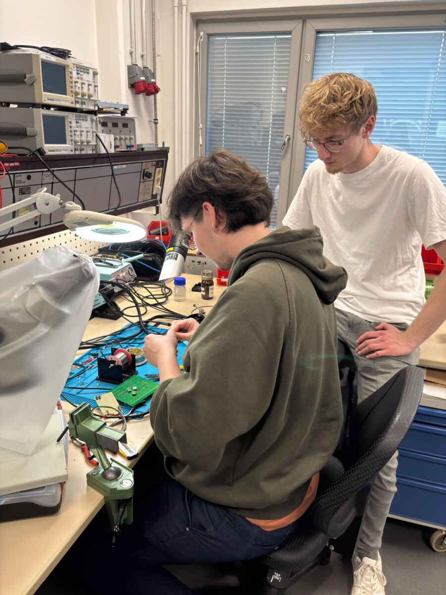 Elektroniikkatyöpisteellä työskentelee kaksi henkilöä. Toinen heistä, joka istuu ja jolla on yllään vihreä huppari, kokoaa piirikomponentteja, kun taas toinen, joka seisoo valkoisessa t-paidassa, seuraa työtä tarkasti. Heidän ympärillään näkyy työkaluja ja laitteita.