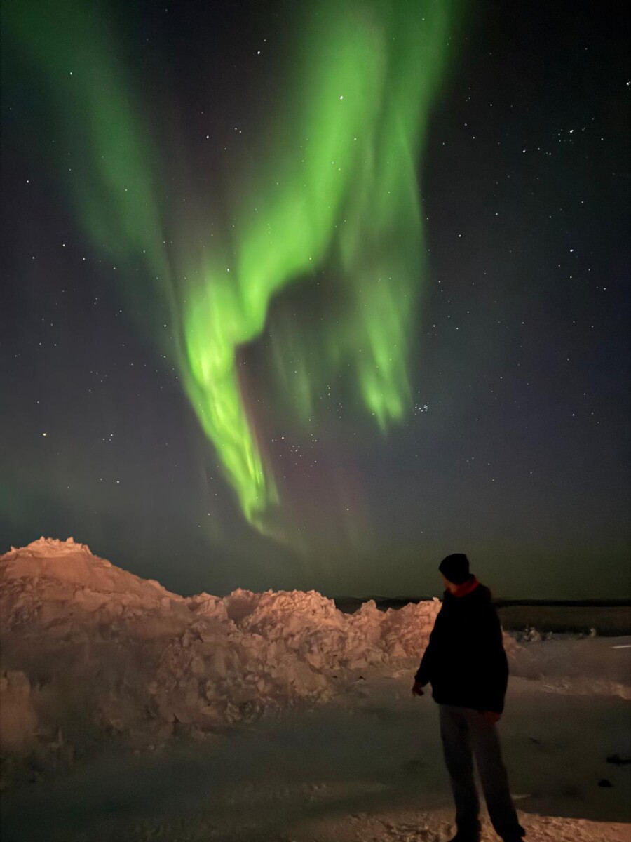 Eräs henkilö seisoo yöllä lumisella maalla ja katselee ylöspäin kirkkaanvihreää revontulia, joka tanssii pimeällä, tähtitaivaalla.