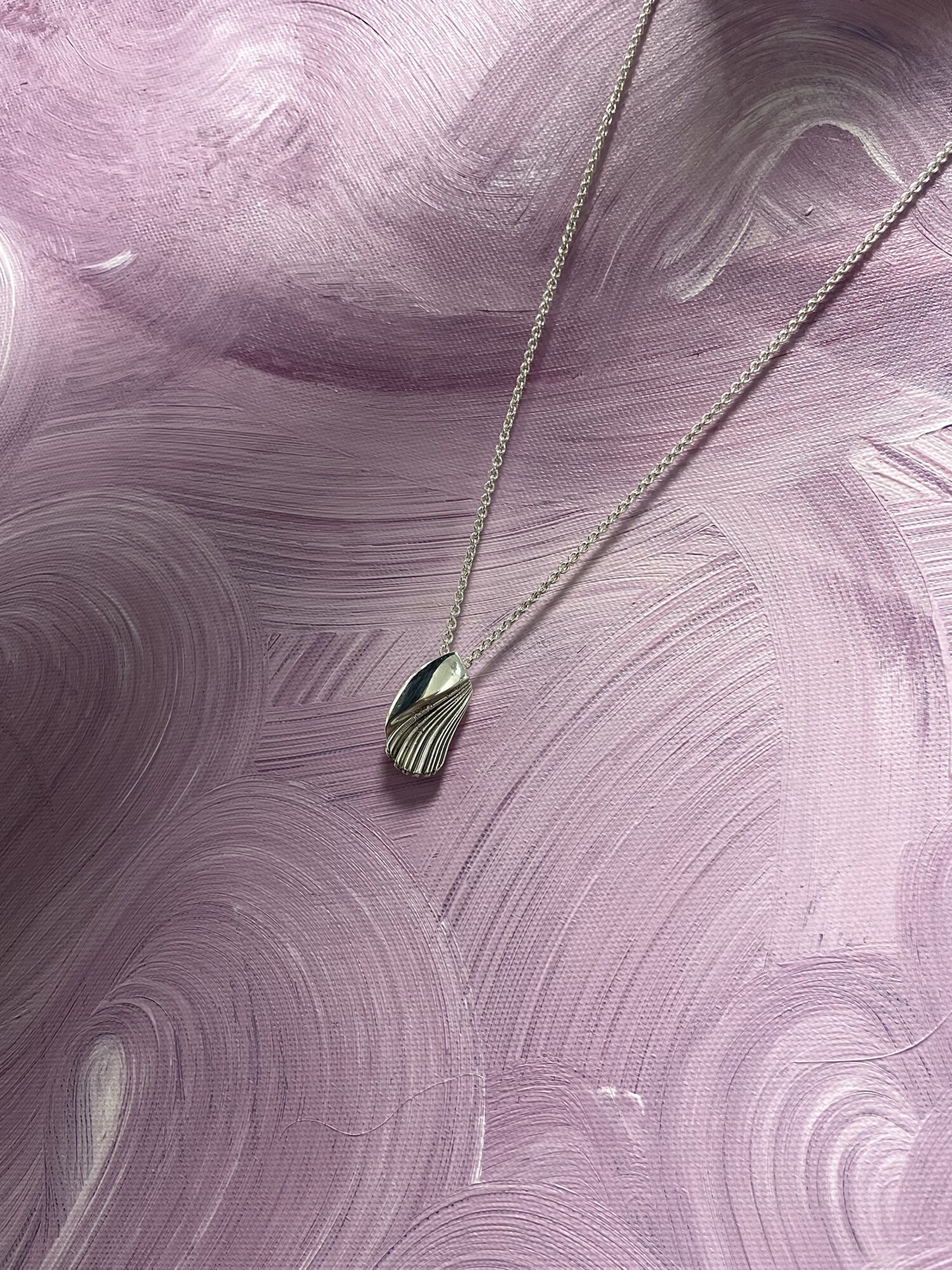 Hopeinen riipuskaulakoru, jossa on harjattu, kuorimainen muotoilu, sijaitsee kuvioidulla, pyörteisellä, pastellivärisellä vaaleanpunaisella ja violetilla maalatulla pohjalla.