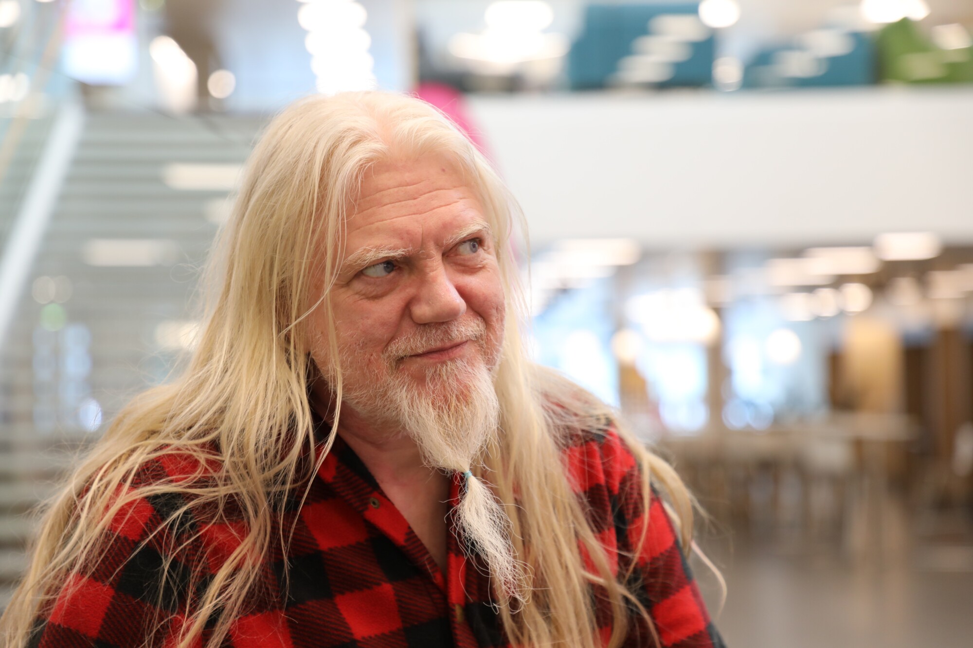 Vanhempi mies, jolla on pitkät valkoiset hiukset ja parta ja joka on pukeutunut punamustaan ruudulliseen paitaan, istuu sisätilassa, jonka taustalla näkyy hämärä valaistus ja moderni sisustus.