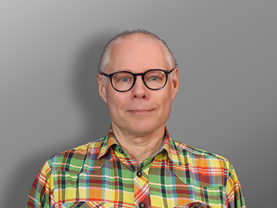 Marko Salmela, Keski-ikäinen mies, jolla on lyhyet harmaat hiukset ja silmälasit, on pukeutunut värikkääseen ruudulliseen paitaan, seisoo harmaan taustan edessä ja katsoo kameraan neutraalilla ilmeellä.