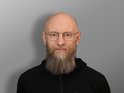 Mustaan huppariin pukeutunut, silmälasipäinen parrakas mies seisoo harmaan taustan edessä ja katsoo neutraalilla ilmeellä suoraan kameraan.