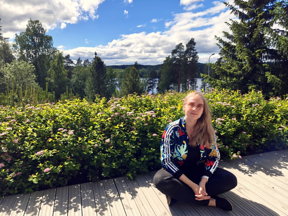 Pitkäkarvainen, vaaleatukkainen nainen, jolla on silmälasit, värikäs takki ja mustat housut, istuu ristikkäin puuterassilla. Hänen takanaan on reheviä vihreitä pensaita, korkeita puita ja kaunis järvi osittain pilvisen taivaan alla.