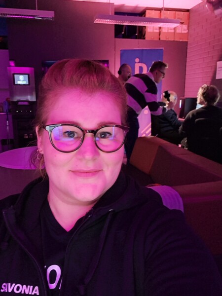 Silmälasit päässä ja hiukset taakse sidottu nainen ottaa selfietä huoneessa, jossa on violetti valaistus, sohvia ja taustalla useita ihmisiä, jotka työskentelevät tai keskustelevat.