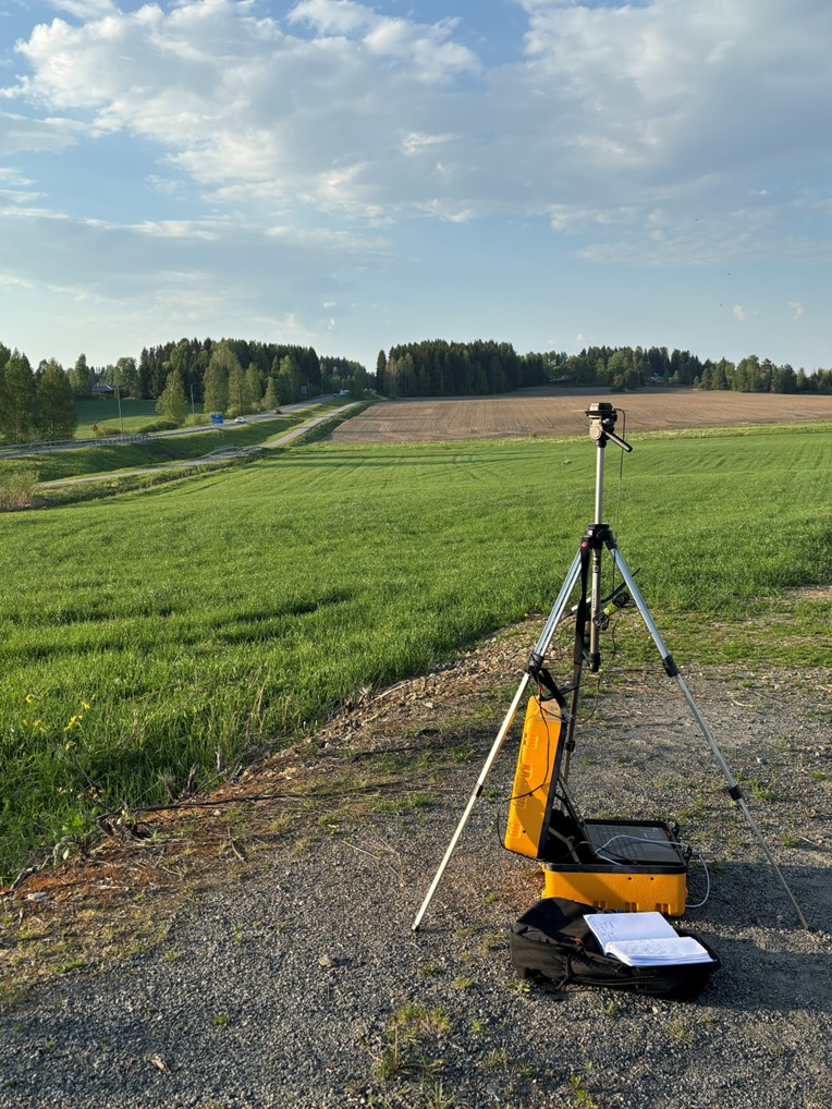 Kolmijalalla seisova kamera on ruohokentällä hiekkatien vieressä, ja maassa on keltainen laukku sekä papereita. Taustalla pellot ja puut ulottuvat osittain pilvisen taivaan alla.