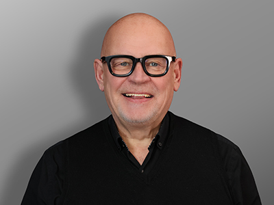 Rauno Tikkanen, Hymyilevä kalju henkilö, jolla on mustakehyksiset silmälasit ja musta paita, seisoo harmaan taustan edessä.