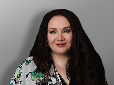Ritva-Liisa Matilainen, Nainen, jolla on pitkät tummat hiukset ja vaaleat silmät, poseeraa harmaan sävytaustan edessä kuvioidussa topissa, jossa on vihreitä ja mustia kukkakuvioita. Hän hymyilee pehmeästi kameralle.