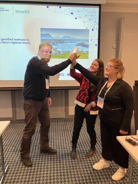 Kolme henkilöä seisoo esitysnäytön edessä, hymyilee ja lyö toisiaan kämmenellä. Kahdella naisella ja yhdellä miehellä on konferenssikortit kaulassaan. Näytöllä näkyy dia, jossa on tekstiä ja kuva rannikkokaupungista.