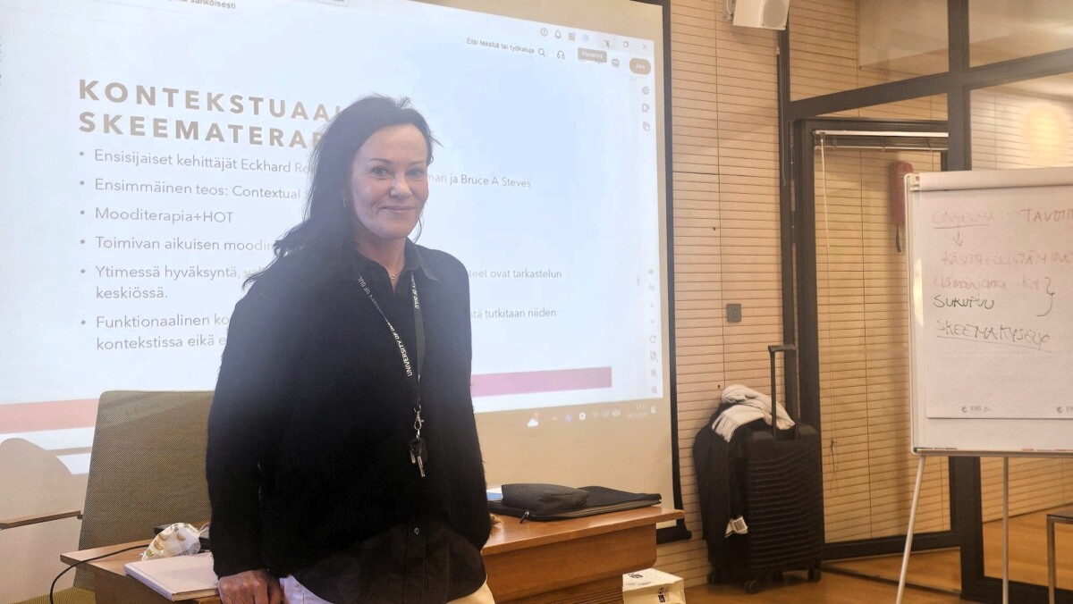 Nainen seisoo ja hymyilee projektorinäytön edessä, jossa näkyy suomenkielinen esitysdia. Oikealla näkyy fläppitaulu, jossa on käsinkirjoitettuja muistiinpanoja, ja huone vaikuttaa luokkahuoneelta tai kokoustilalta.