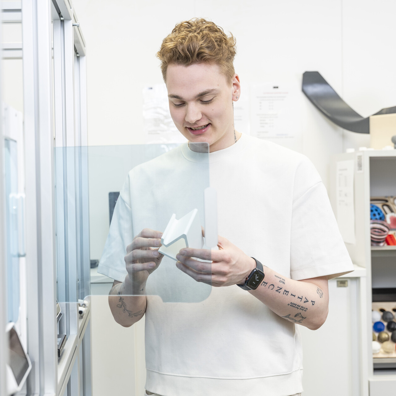Lyhyet kiharat hiukset omaava nuori tutkii valkoista 3D-tulostettua esinettä modernissa laboratoriossa, jota ympäröivät hyllyt ja laitteet.
